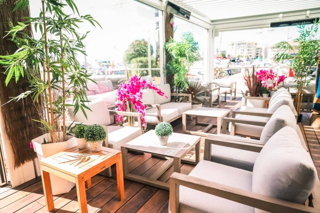 Terrasse ensoleillée avec meubles confortables, tables boisées, plantes et fleurs roses éclatantes.
