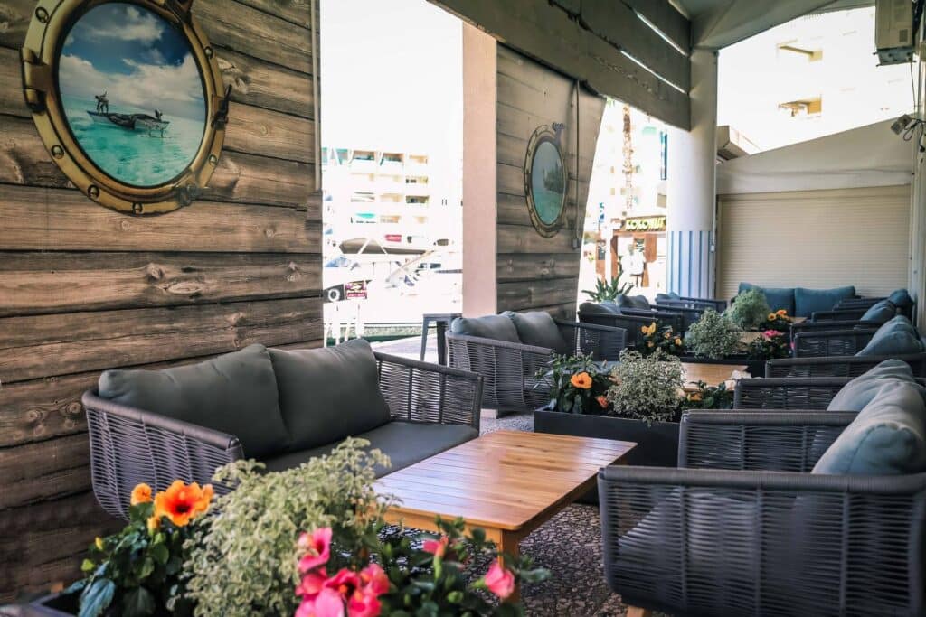 Terrasse avec canapés en osier gris, tables en bois, plantes en pot et vues pittoresques sur le port.