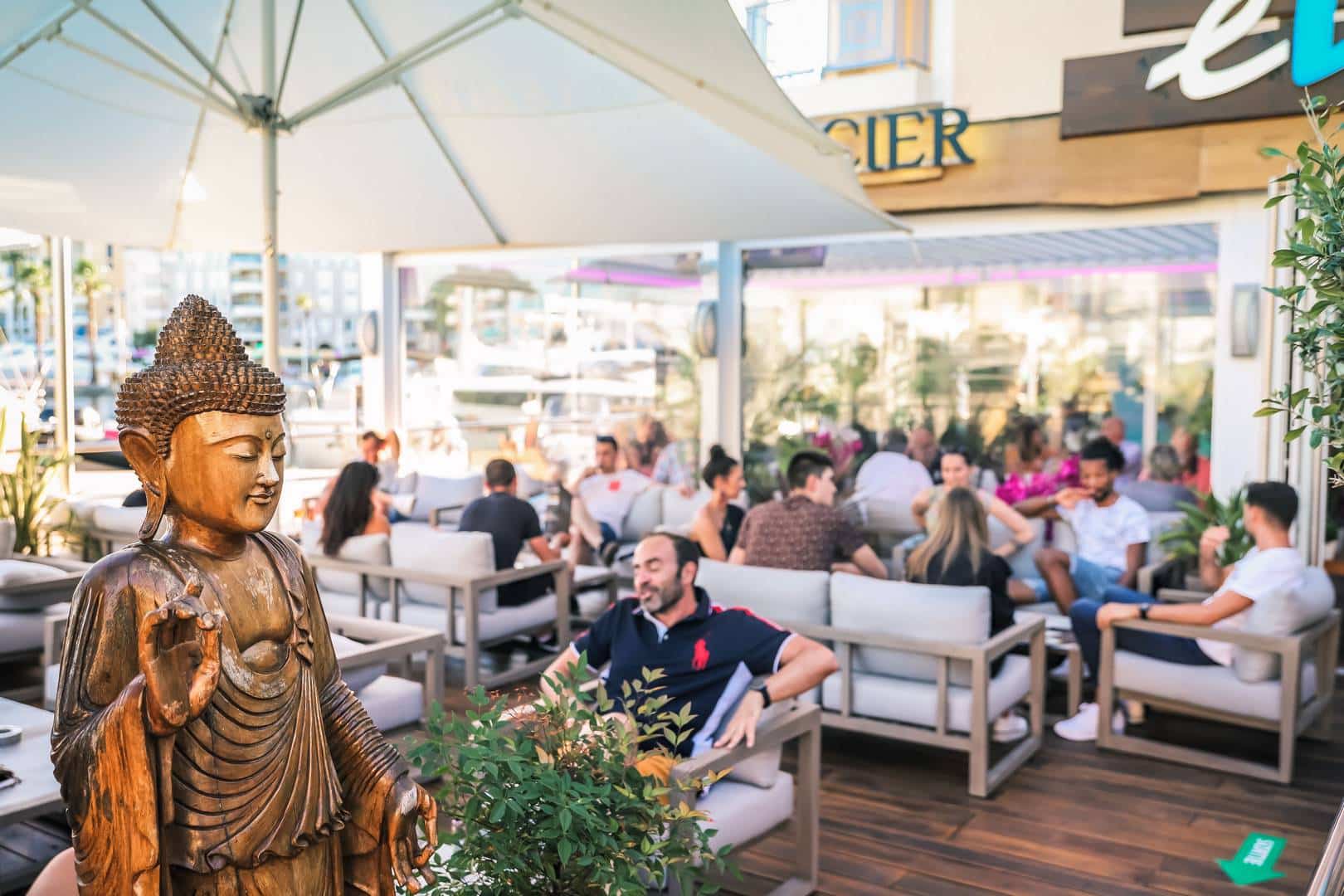 Des clients discutent en terrasse avec une ambiance accueillante, ombragée par des parasols et verdoyante.
