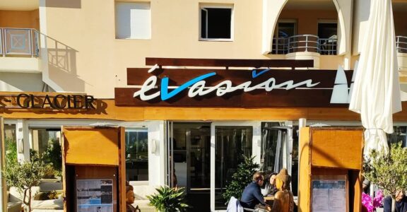 Entrée de L'Évasion, Fréjus : terrasse extérieure, présentoirs à menus en bois, parasol blanc. Ambiance délicieuse.